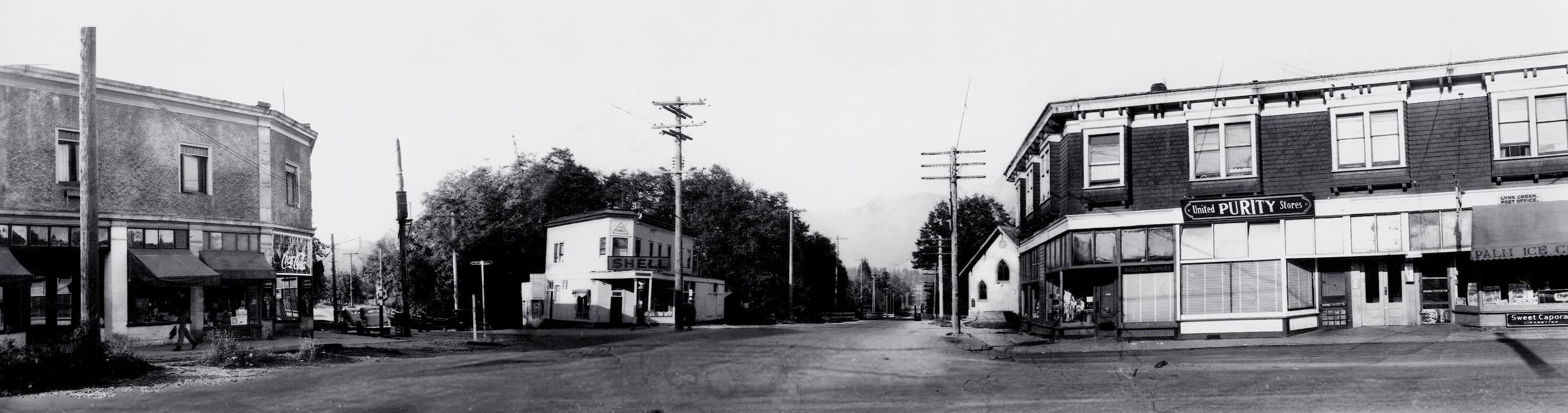 ca. 1950s. Panoramic image along Lynn Valley Road, across from Mountain Hwy. Buildings depicted from left to right: the Brier Block; the Triangle block; the Fromme block. Lynn Valley United Church can be seen behind the Fromme block. This image was taken after the streetcar lines had been removed.